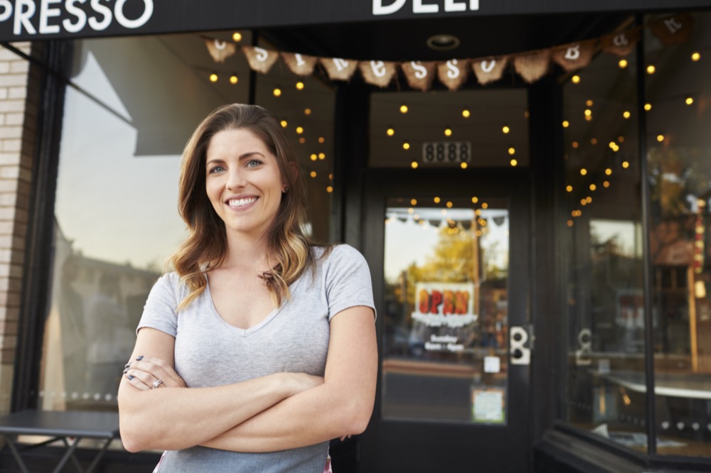Smiling female business owner standing with arms crossed outside her boutique cafe entrance, representing web design and SEO for local entrepreneurs.