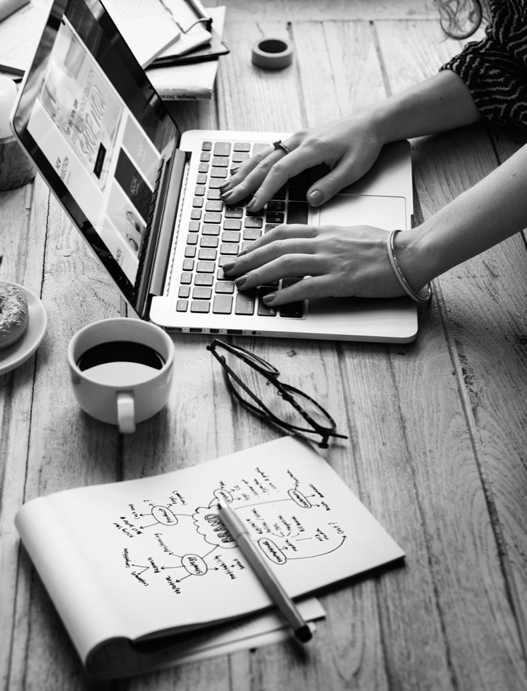 Overhead black-and-white photo of a freelancer working on a laptop next to a coffee cup, reading glasses, and a notebook with a hand-drawn brand strategy mind map.