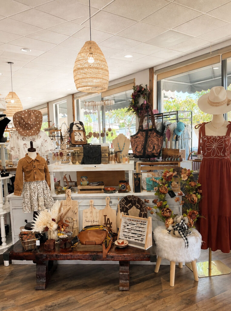 Boho chic boutique interior with displays of women's apparel, rust-colored dress on a mannequin, leather handbags, wide-brim hats, and wooden cutting boards.