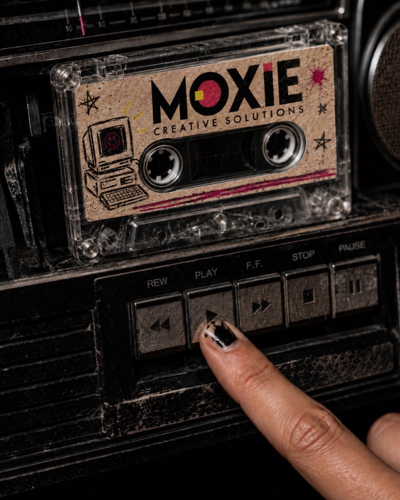 A close-up of a vintage cassette player with a clear audio cassette inside. The cassette label features the text "MOXIE CREATIVE SOLUTIONS" alongside retro desktop computer and star doodles. A hand with chipped black nail polish is shown pressing the metal "PLAY" button on the cassette deck.